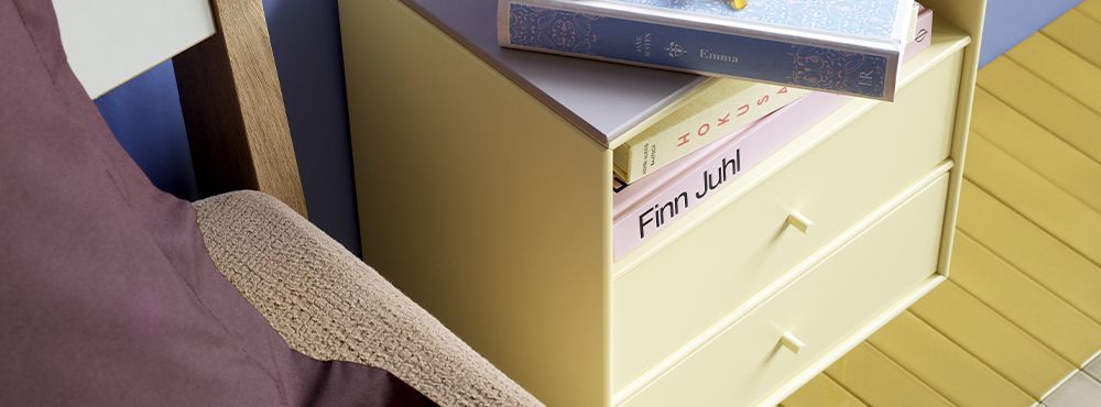 En lille skænk i lys gul med to skuffer og et par bøger ovenpå.