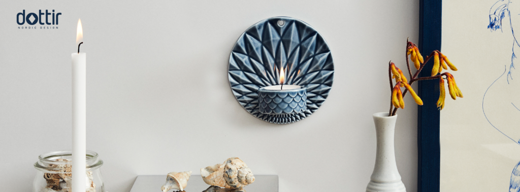 Der hænger en keramiktallerken på væggen med en tilhørende lille skål til et fyrfadslys, som er tændt. På bordet står et kronelys tændt og en hvid vase med en blomst i.