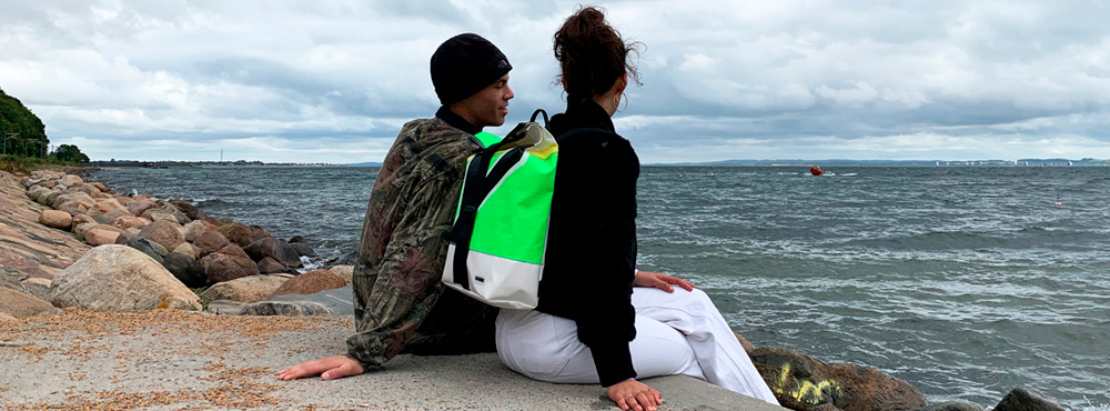 To mennesker sidder ved på en klippe ved havet. Kvinden har en rygsæk på.